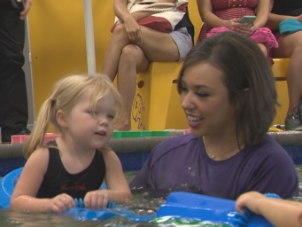 A female swim instructor in purple shirt helps a girl learn to swim while she holds on to a floaty