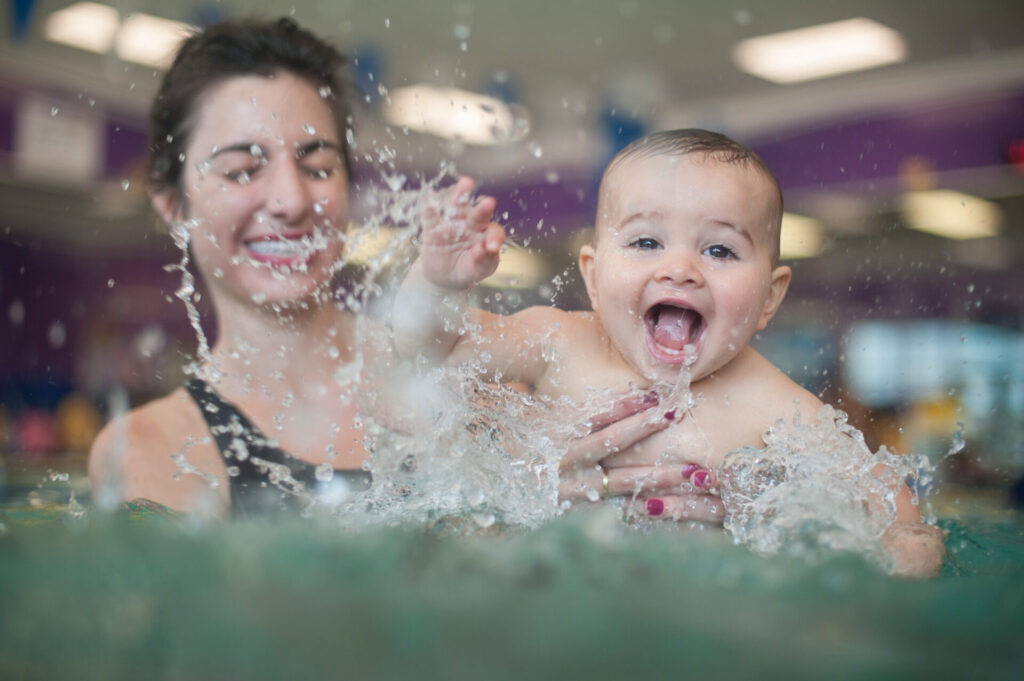 Mother holding a smiling baby boy in front of her as he splashes in the pool